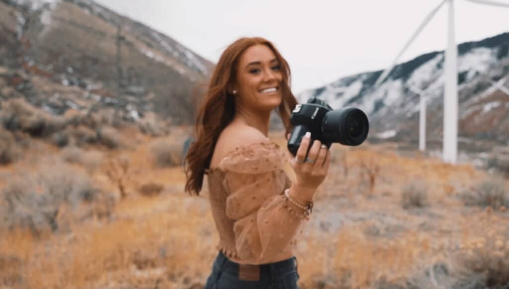 TIktok Videographer girl holding camera in mountains with windmills in the background really pretty girl red head holding camera in field with snow in background and mountains. top 10 best cameras 2025 dslr mirrorless teaches how to stay trending on social media 2024